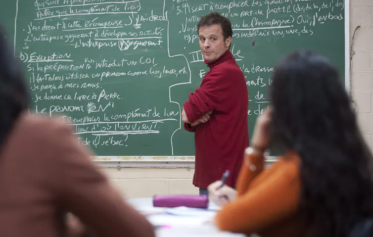 A middle-aged white man stands in front of a chalkboard at the front of a classroom