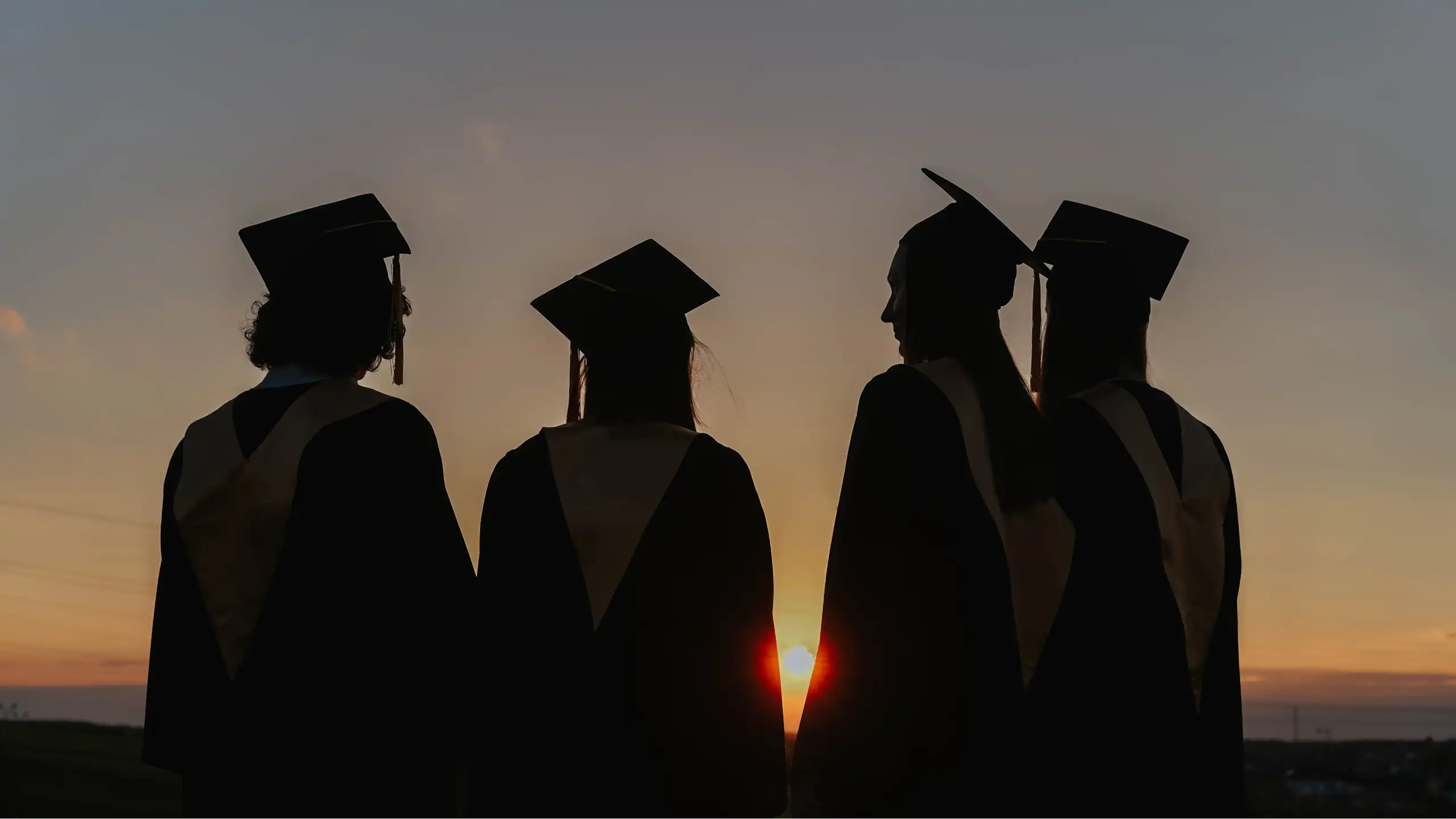 The silhouettes of four young people in graduation robes, looking at a sun set