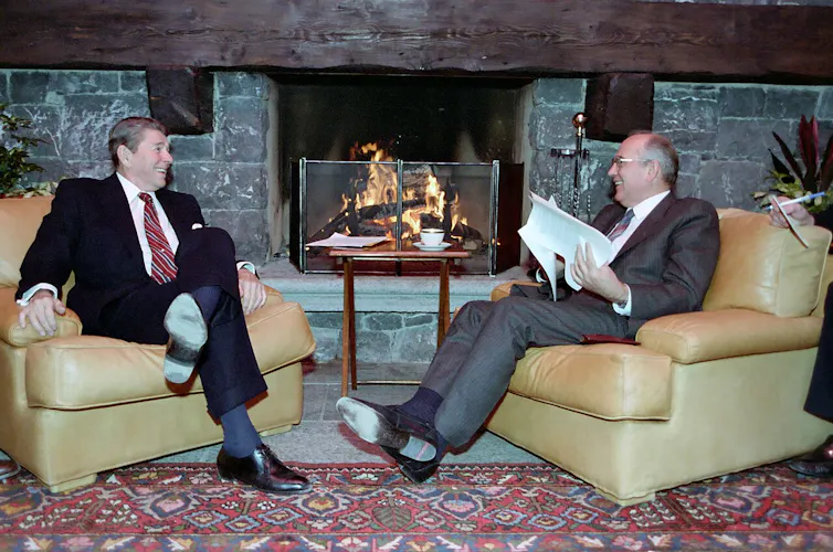 Ronald Reagan and Mikhail Gorbachev sit smiling at each other jn front of a log fire.