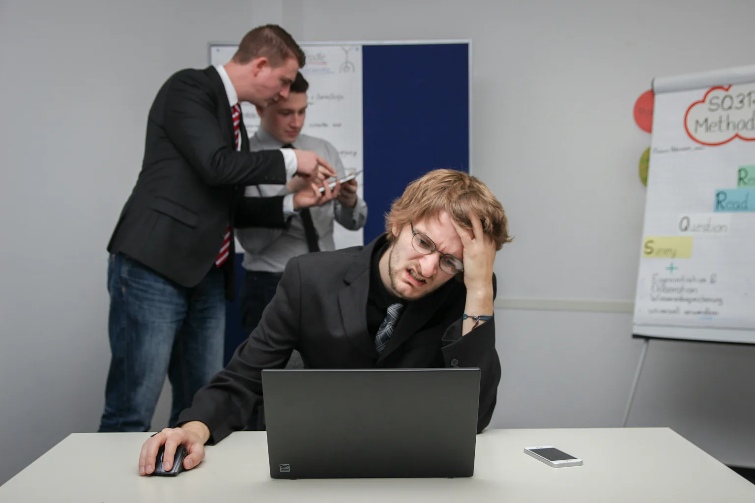 A young man with glasses looks confused and exasperated as he sits in front of a computer, while two colleagues discuss something behind him.