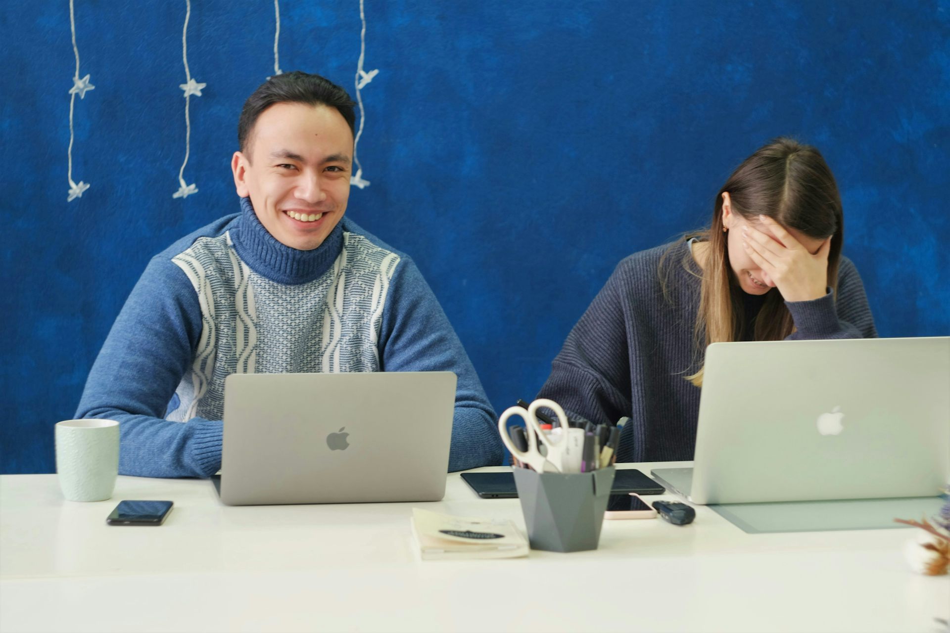 One young worker in a white and blue sweater grins while his colleague, a woman with long hair, puts her head in her left hand.