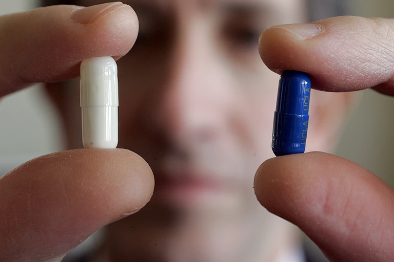 Man's face blurry in background pinching a white pill with one hand and a blue pill with the other
