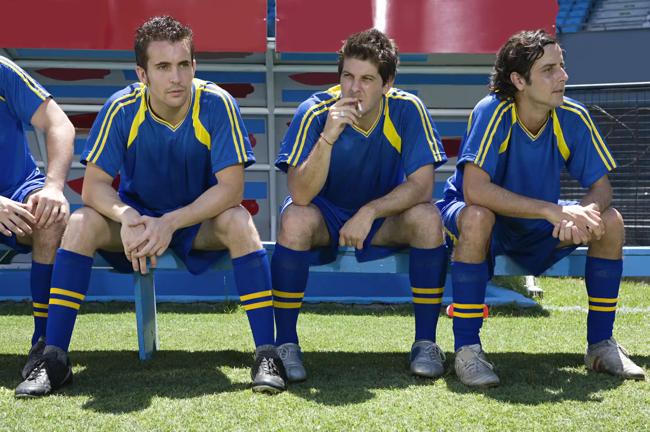 Three men in soccer uniforms sit on a bench, while the one in the middle smokes a cigarette.