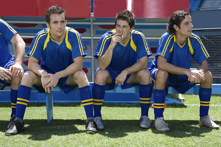 Three men in soccer uniforms sit on a bench, while the one in the middle smokes a cigarette.