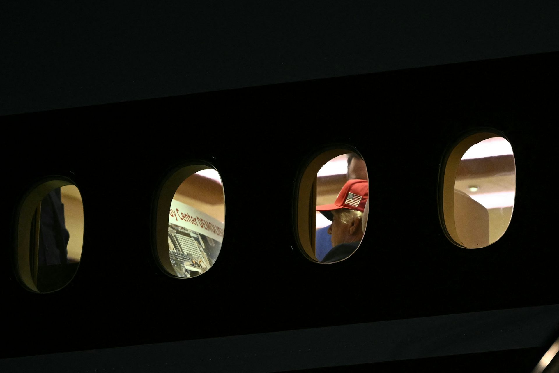 A man in a baseball cap is seen aboard an airplane, apparently reading the words 'Kennedy Center demolished.'