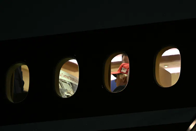 A man in a baseball cap is seen aboard an airplane, apparently reading the words 'Kennedy Center demolished.'