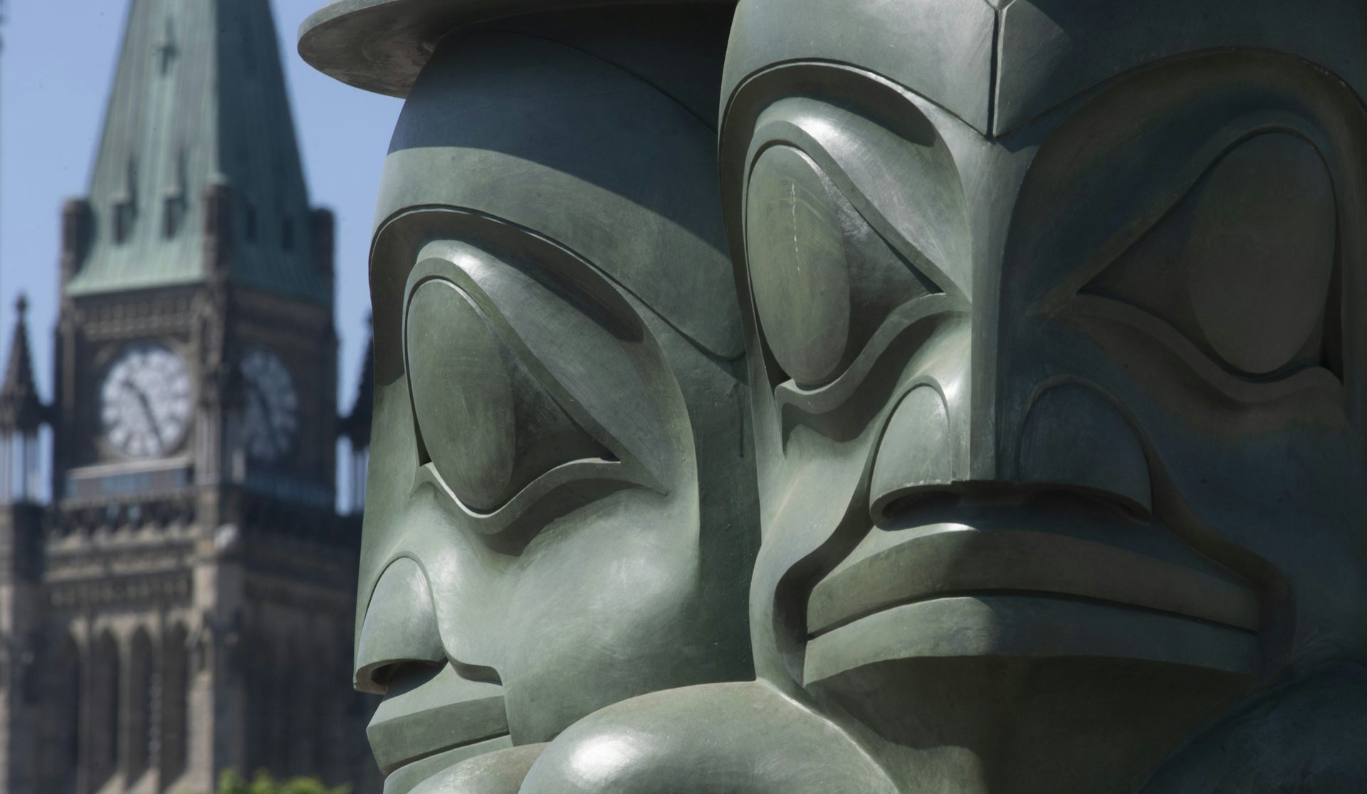 A green-toned statue of watchful figures in front of a buildings with a peaked roof and clock.