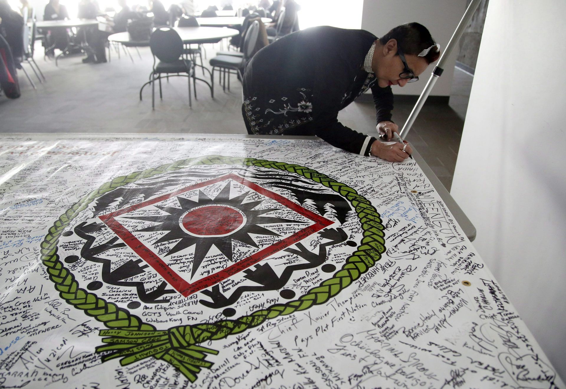 A person holds a marker and signs a banner laid on a table showing a circle with a braided texture and figures inside it encircling a red circle surrounded by a square.