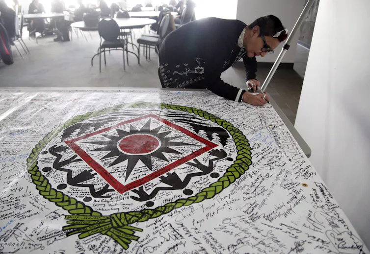 A person holds a marker and signs a banner laid on a table showing a circle with a braided texture and figures inside it encircling a red circle surrounded by a square.