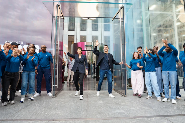 Two people waving as they walk out of a glass door, with small crowds of people on either side applauding.