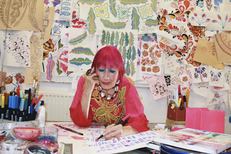 An older lady with pink hair and fabulous clothes sitting a desk sketching.