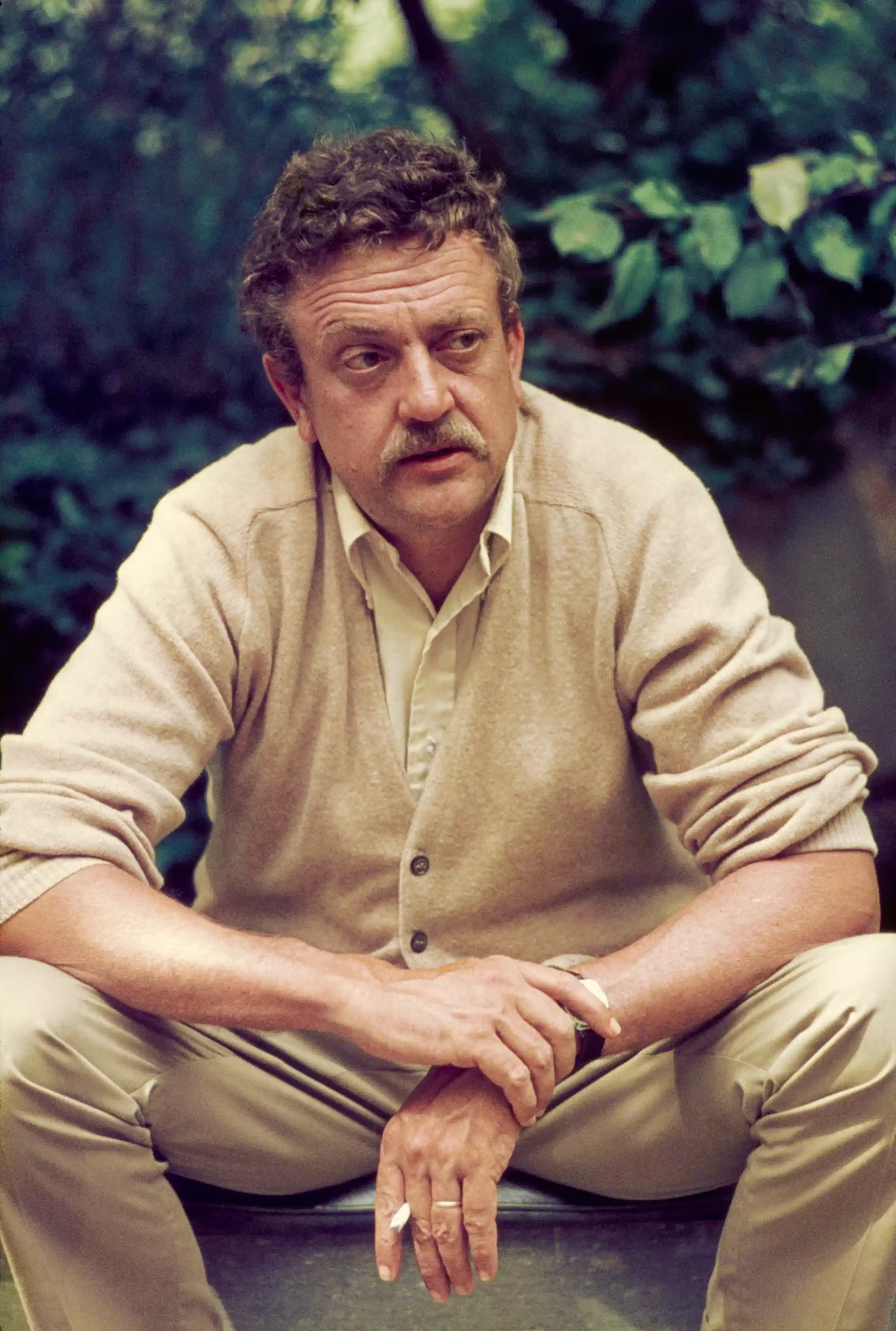 Older man with mustache and curly hair seated and hunched over, resting his arms on his knees and cradling a cigarette in one hand.