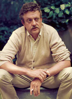 Older man with mustache and curly hair seated and hunched over, resting his arms on his knees and cradling a cigarette in one hand.
