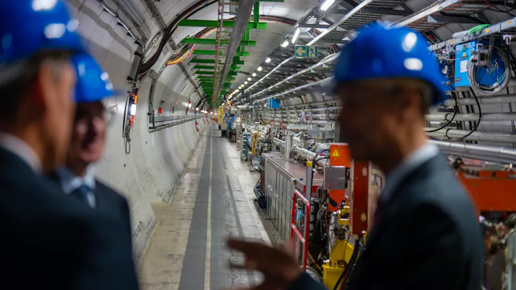 A large, long metal tube in a warehouse-like hallway. Three scientists wearing hard hats are speaking in a group in the foreground.