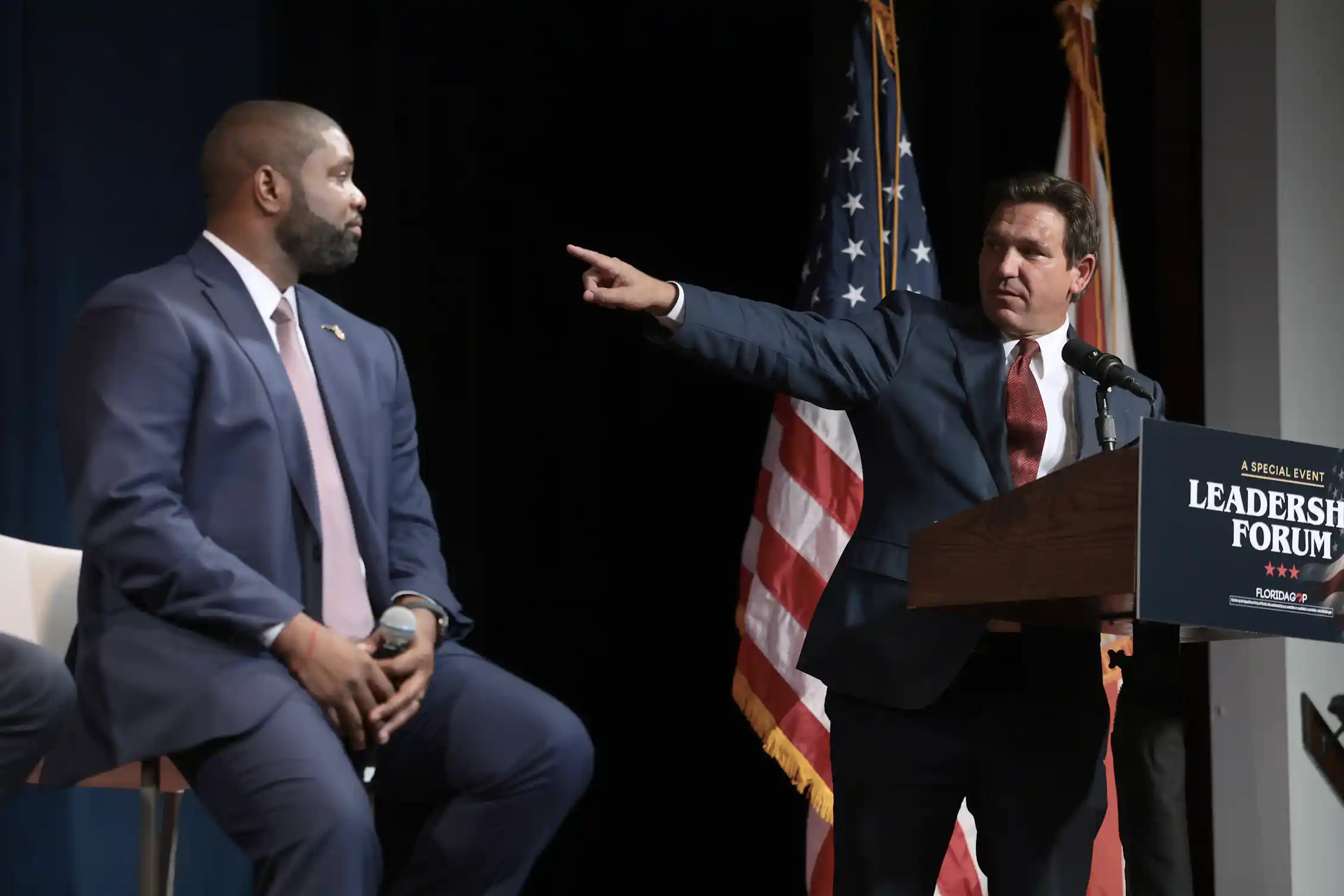 Middle-aged white man wearing suit standing at a podium points at a middle-aged Black man in a suit seated to his right.