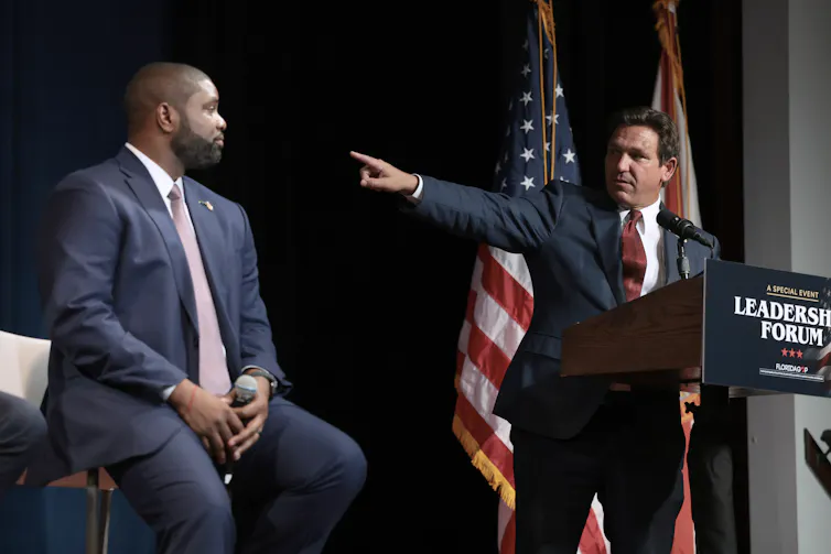 Middle-aged white man wearing suit standing at a podium points at a middle-aged Black man in a suit seated to his right.