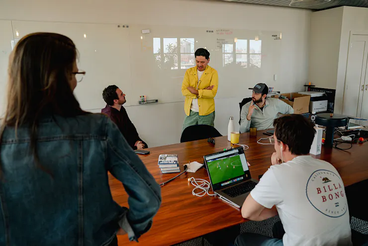 Man wearing yellow shirt folds his arms while speaking and standing in front of a whiteboard as three male and one female staffers look on.