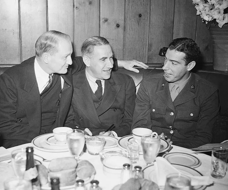 Black-and-white photo of a young man in a military uniform chatting with two older men wearing suits while seated at a dinner table.