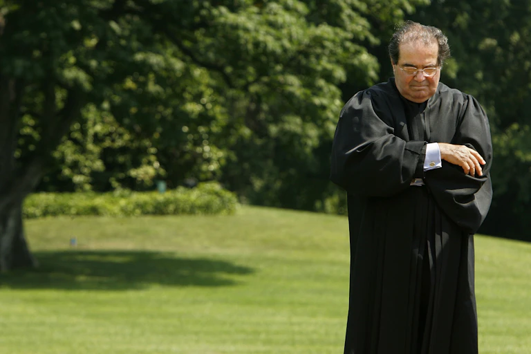 A dark-haired man with glasses, standing on a lawn and wearing a black robe.