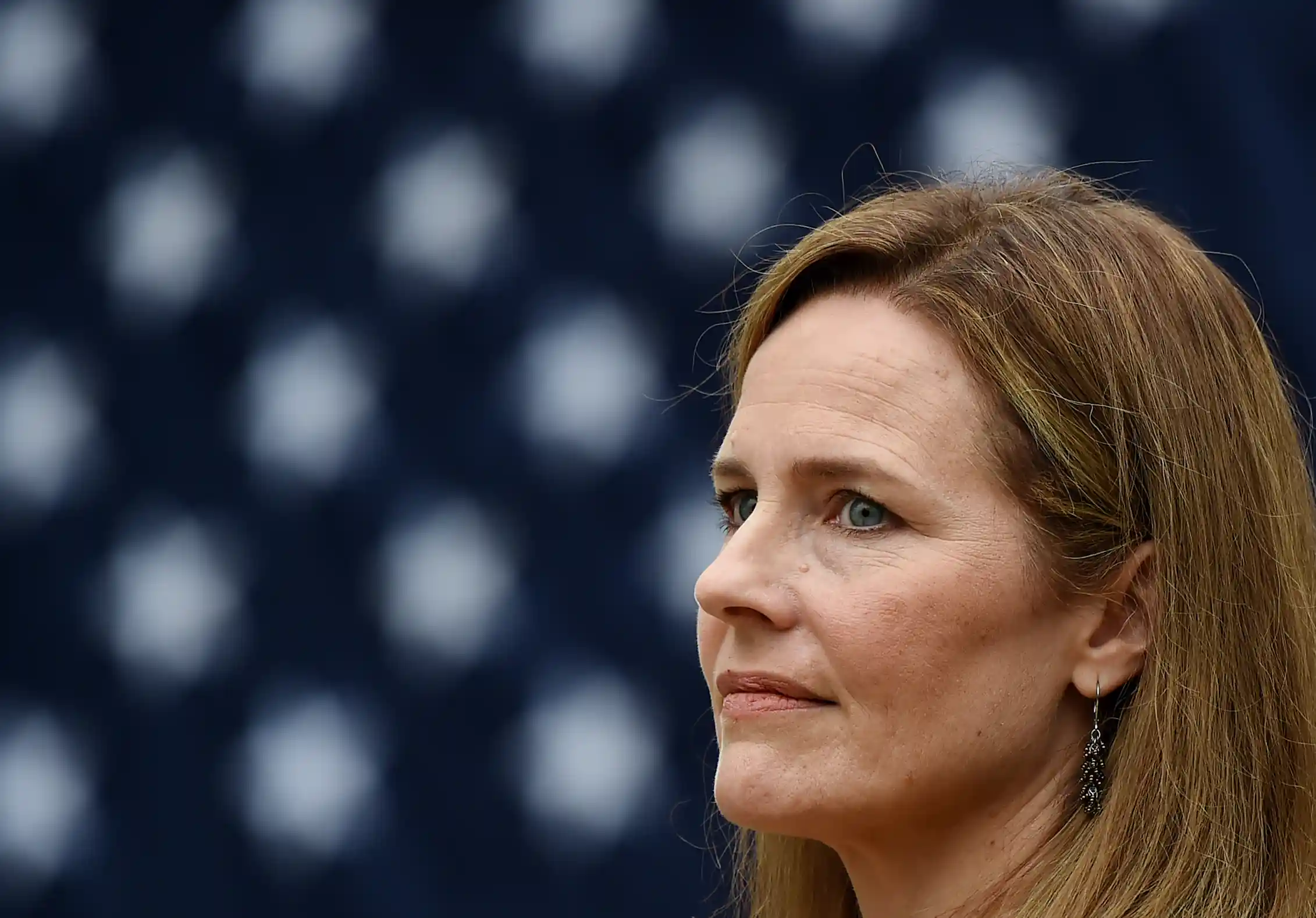 A woman with brown hair looking pensive, with an American flag behind her.