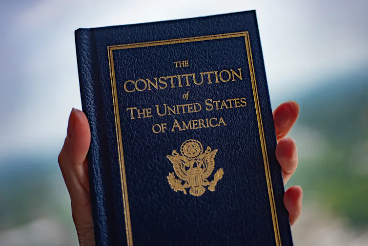 A blue book with the gold title 'The Constitution of The United States of America' being held in someone's hand.