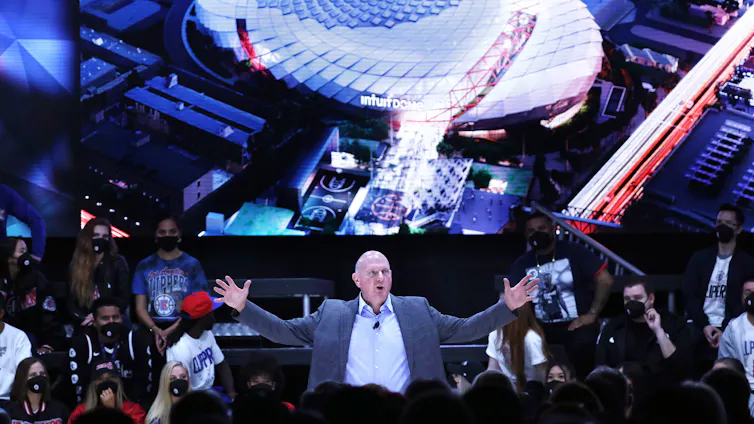 Middle-aged bald man wearing a suit raises his arms while speaking at a podium surrounded by fans.