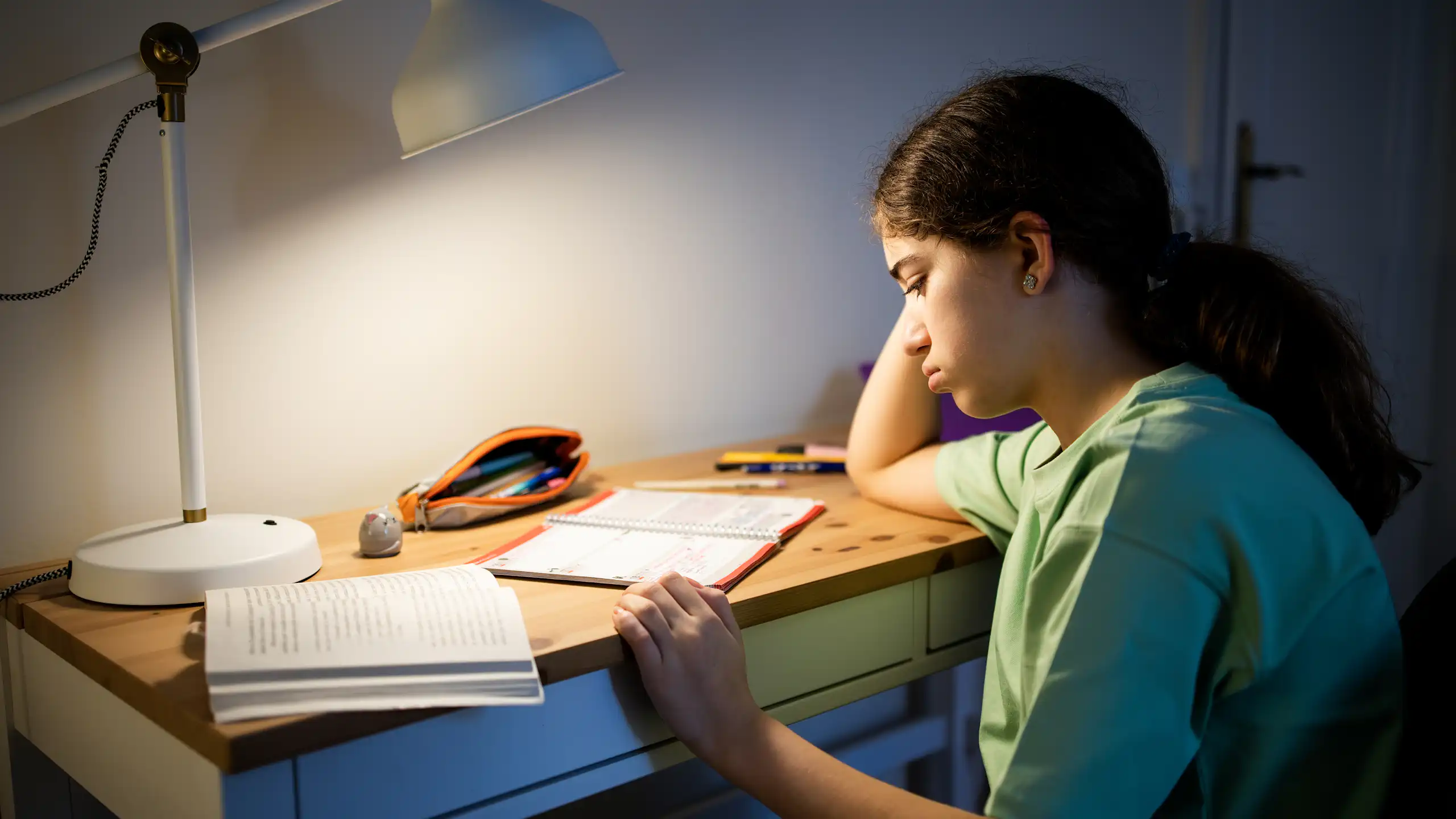Female student leaning on her arm, sitting at a desk and studying under a warm desk lamp in a dark room.