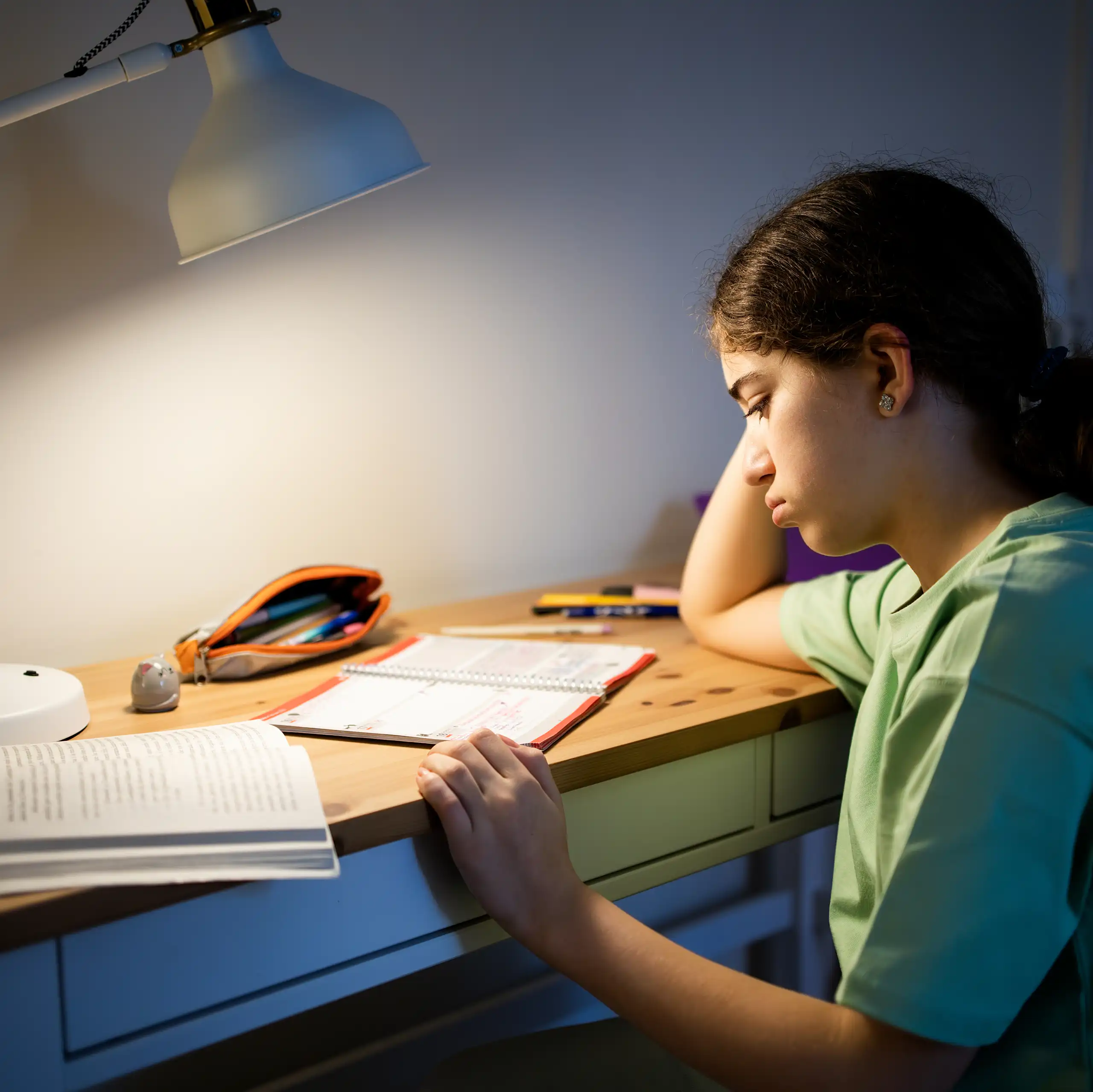 Female student leaning on her arm, sitting at a desk and studying under a warm desk lamp in a dark room.