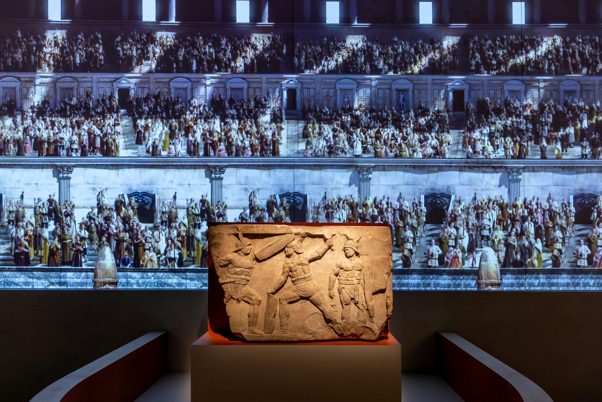 A relief showing fighting gladiators, on a plinth, with digital projections on the gallery wall behind.