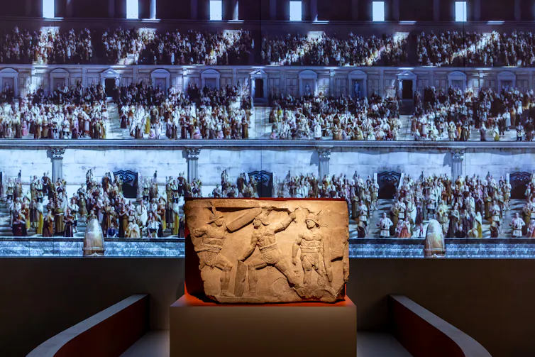 A relief showing fighting gladiators, on a plinth, with digital projections on the gallery wall behind.