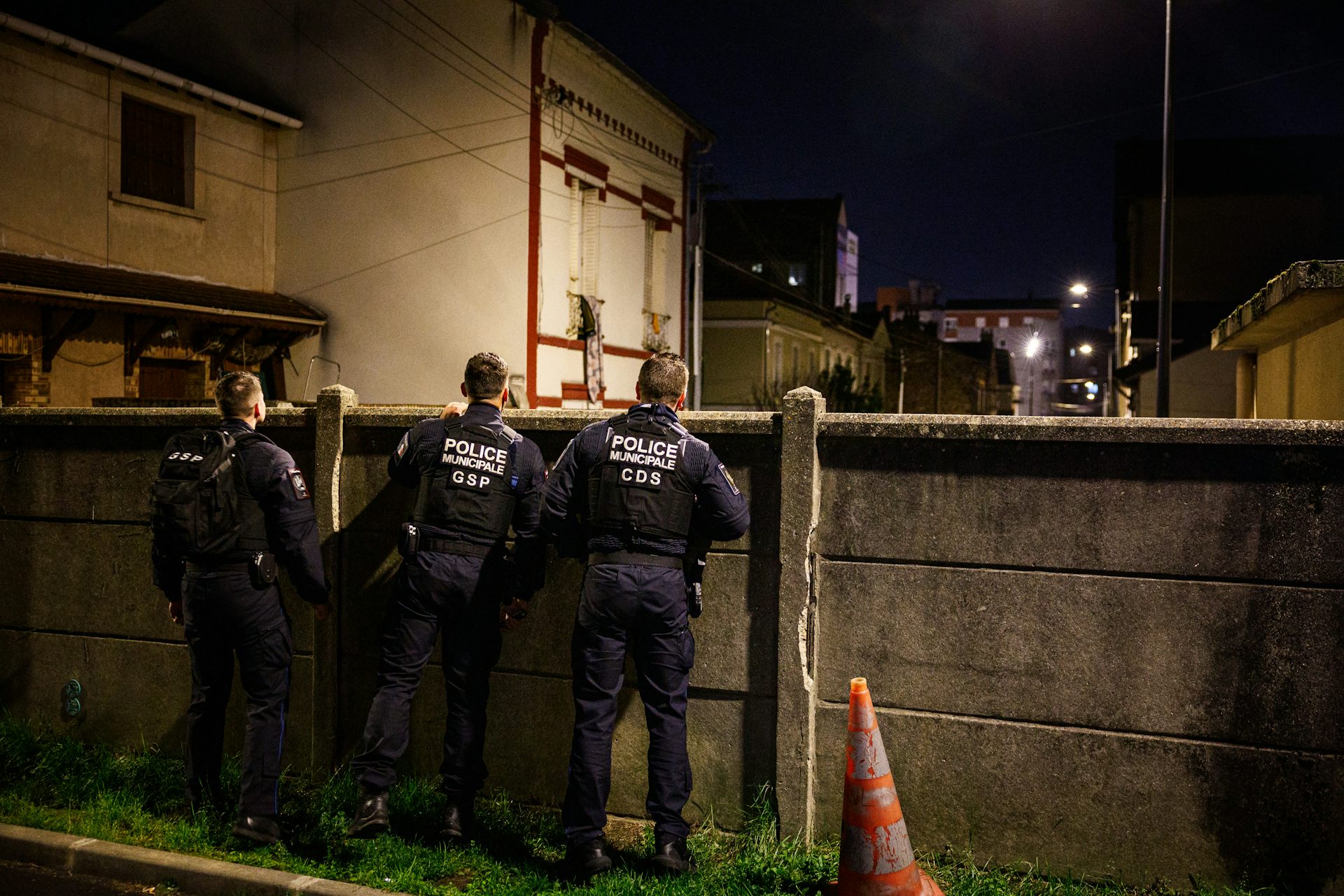 Policiers municipaux vus de dos, surveillant une rue par delà une palissade en béton, la nuit.