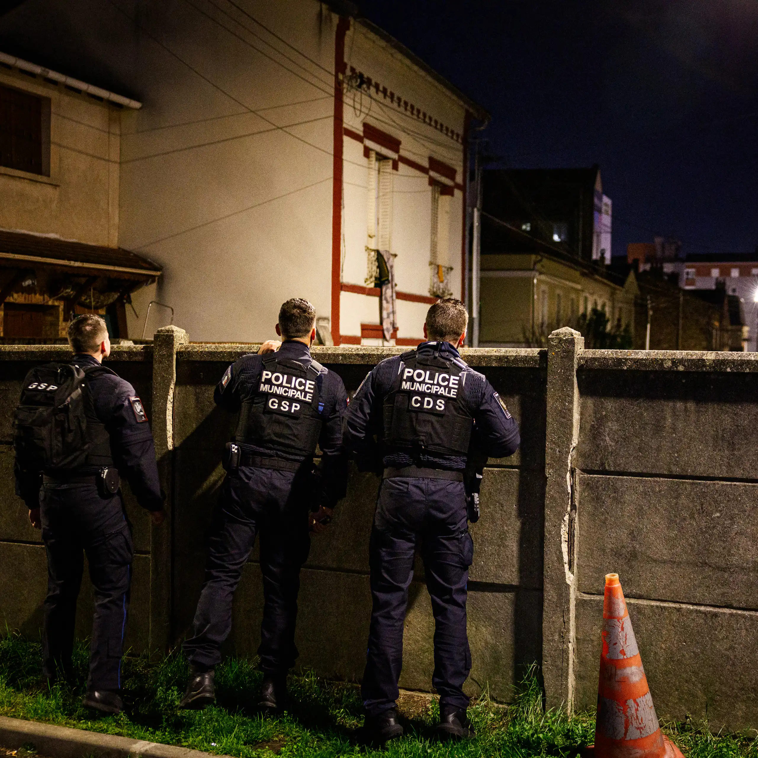 Policiers municipaux vus de dos, surveillant une rue par delà une palissade en béton, la nuit.