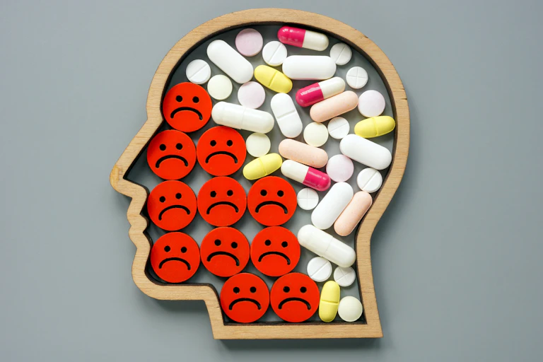 Wooden outline of a human head filled with tablets, capsules and red discs displaying sad faces, on a grey background.