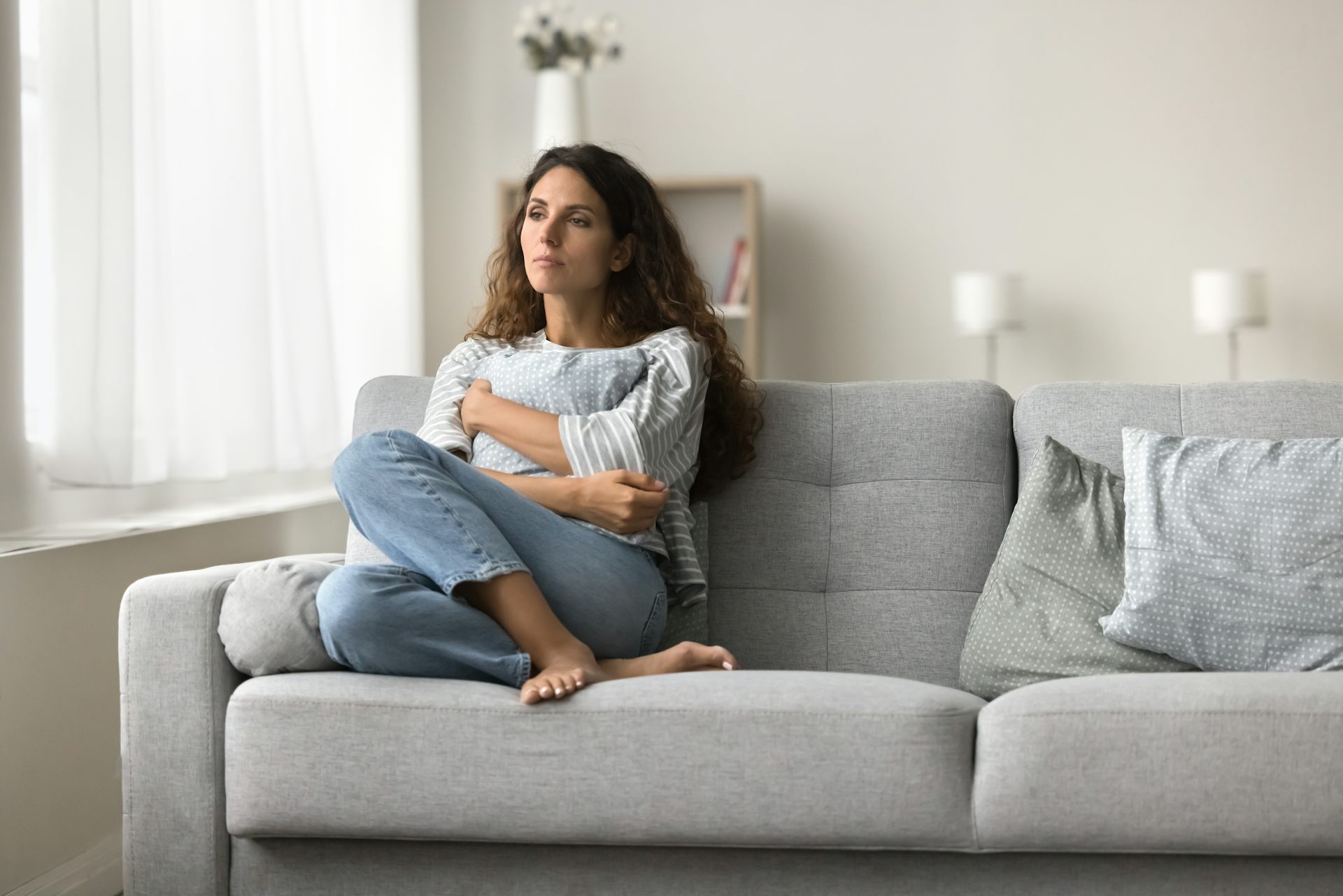 A woman sits on a couch holdings a pillow to her stomach. She looks pensive.