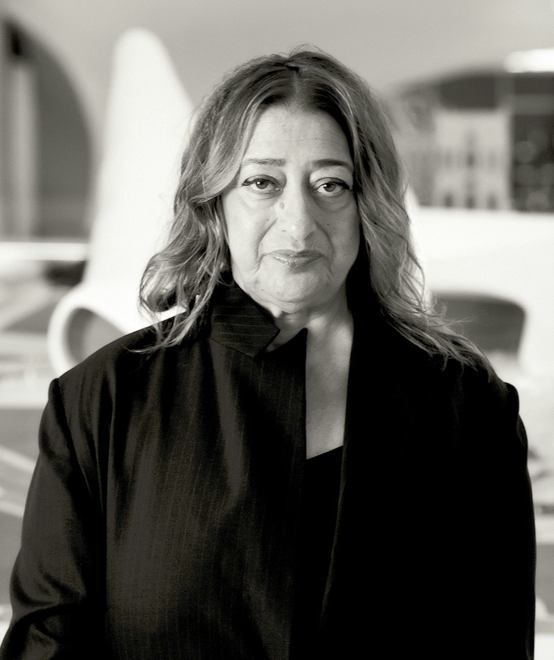 A woman standing in front of a vast curvaceous white building wearing a black suit.