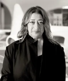 A woman standing in front of a vast curvaceous white building wearing a black suit.
