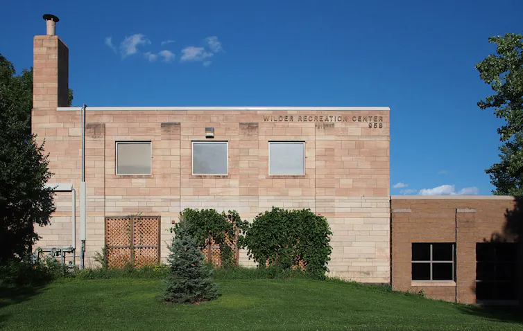 A tan stone building sits on a plot of land with a green yard and a few trees.