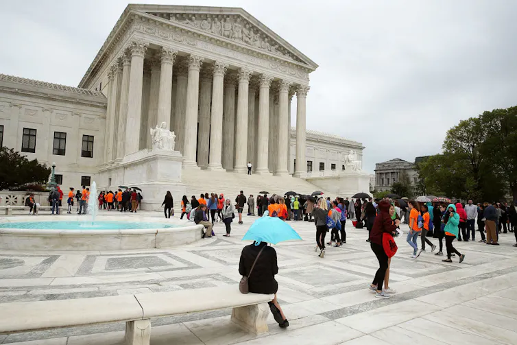 A few dozen people with umbrellas stand in a plaza outside a building with tall marble pillars.