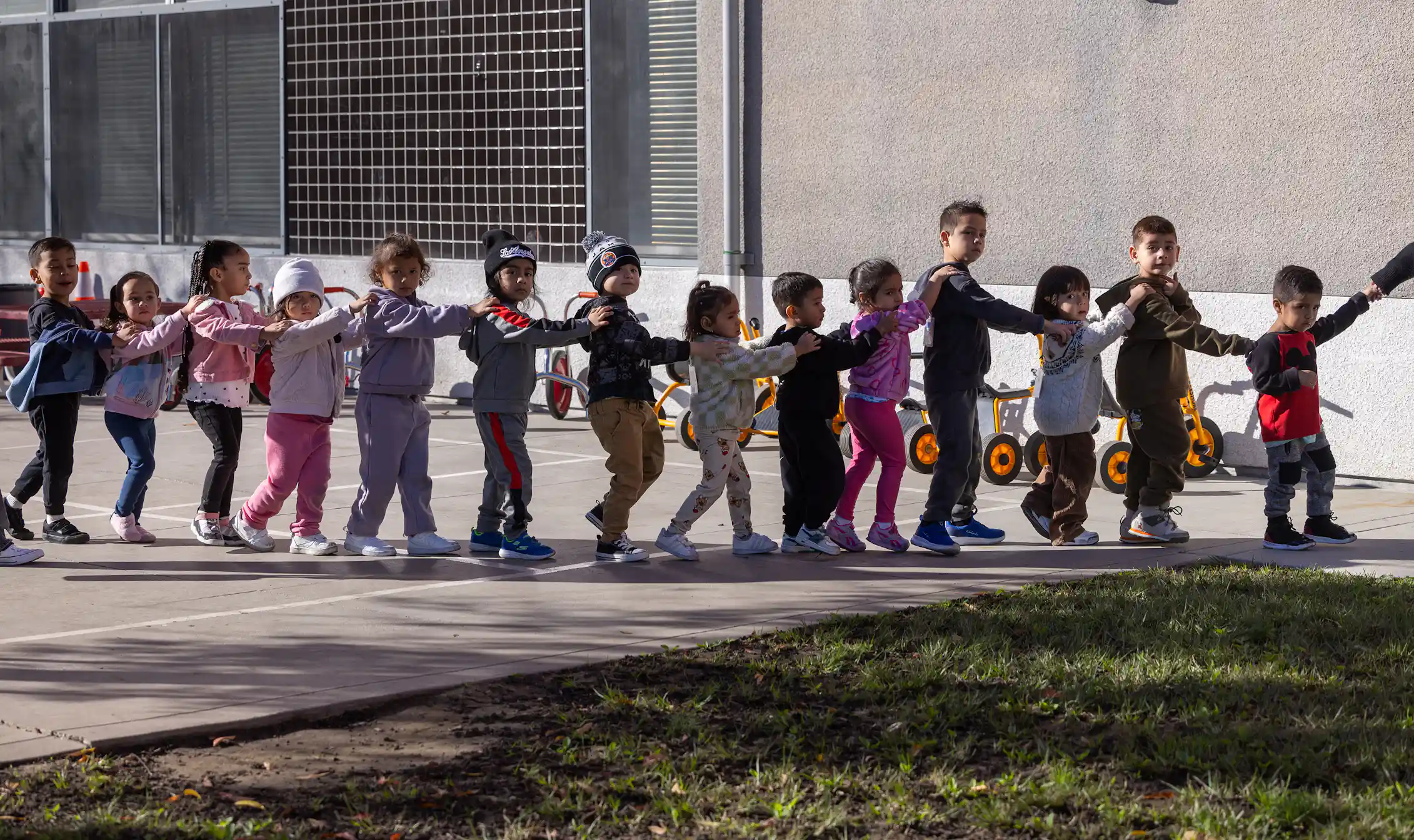 A group of young children walk in a line, with their hands on the shoulders of the child in front of them.