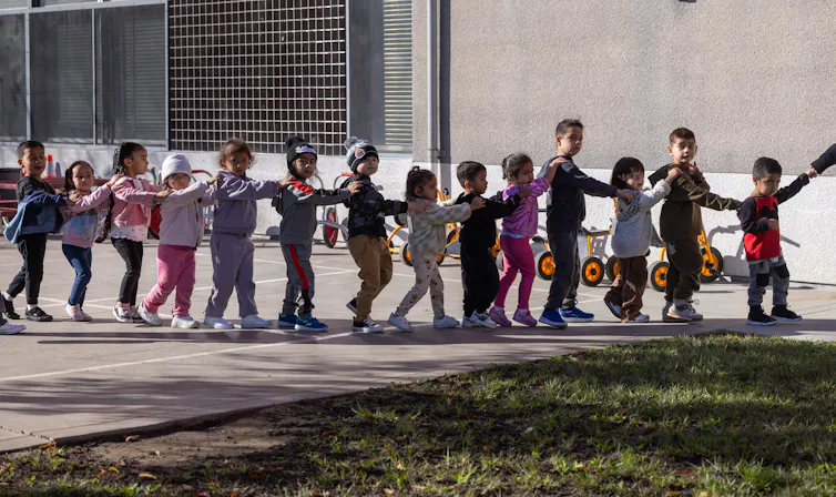 A group of young children walk in a line, with their hands on the shoulders of the child in front of them.