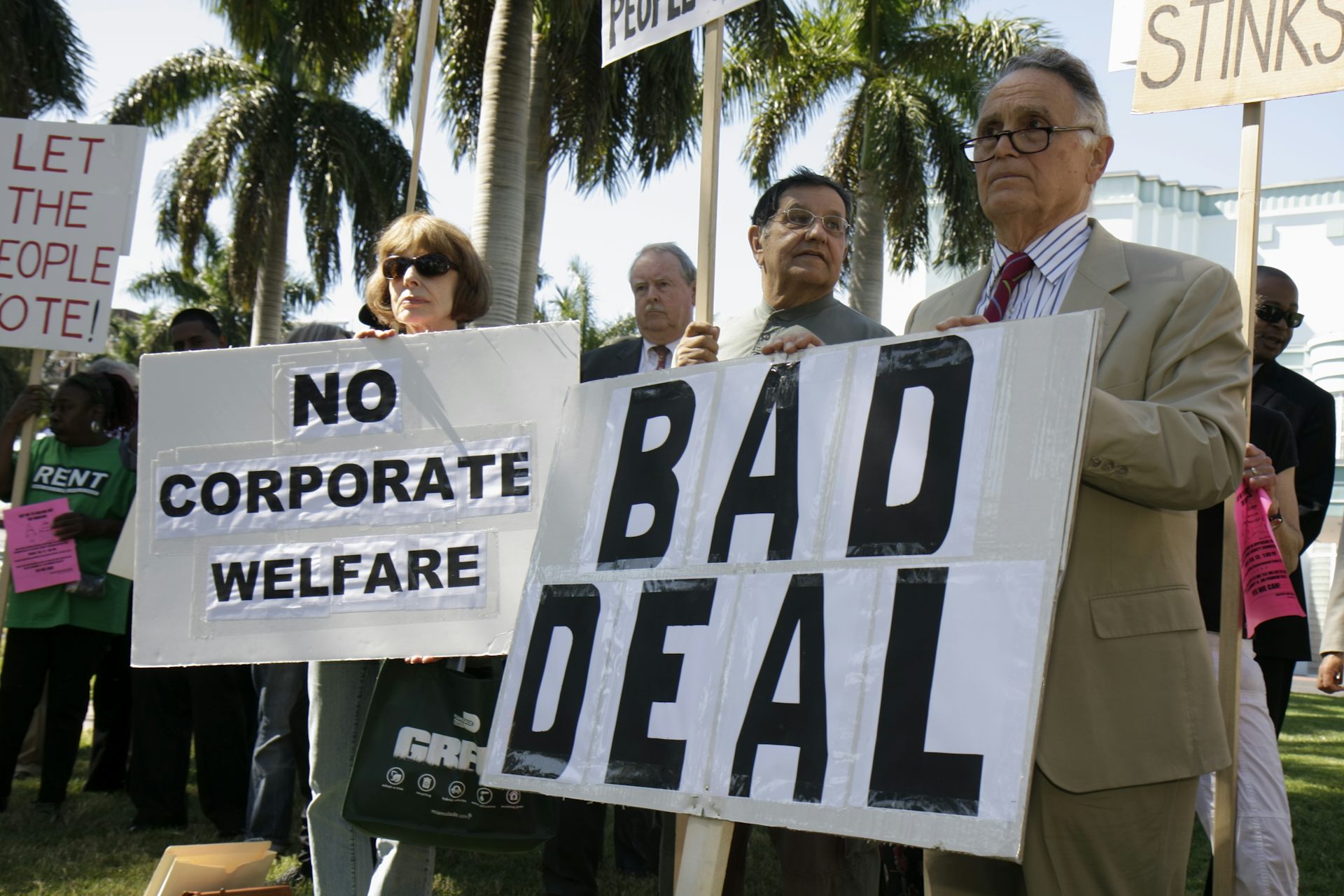 Protesters in Miami, with palm trees behind them, hold signs opposing the Marlins baseball stadium construction deal, citing taxpayer cost.