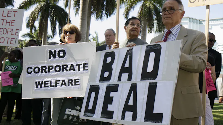 Protesters in Miami, with palm trees behind them, hold signs opposing the Marlins baseball stadium construction deal, citing taxpayer cost.