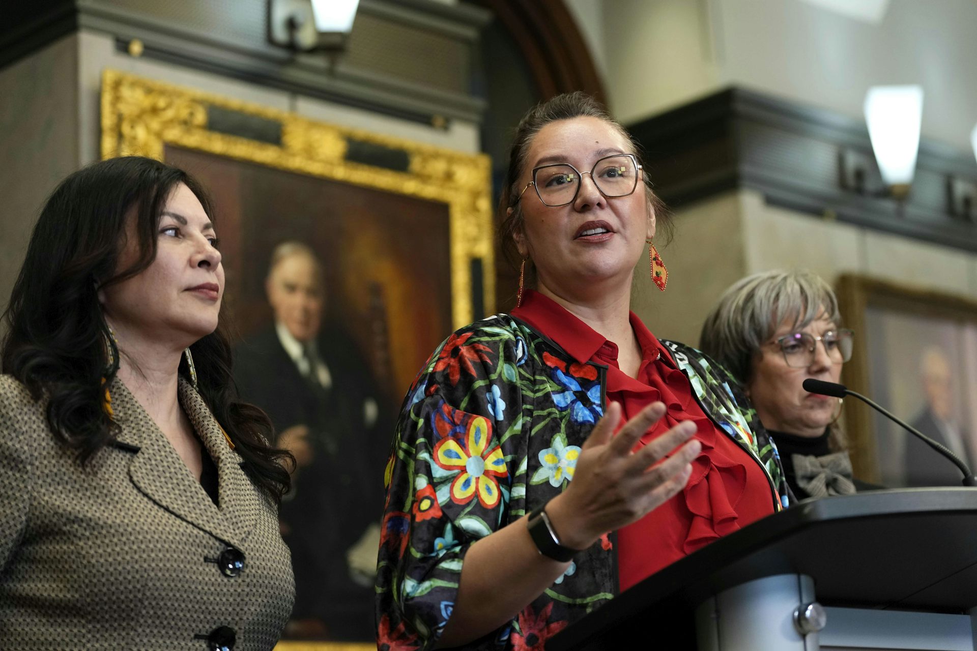 Three women standing at a podium with one in the center leading and speaking at a press conference.