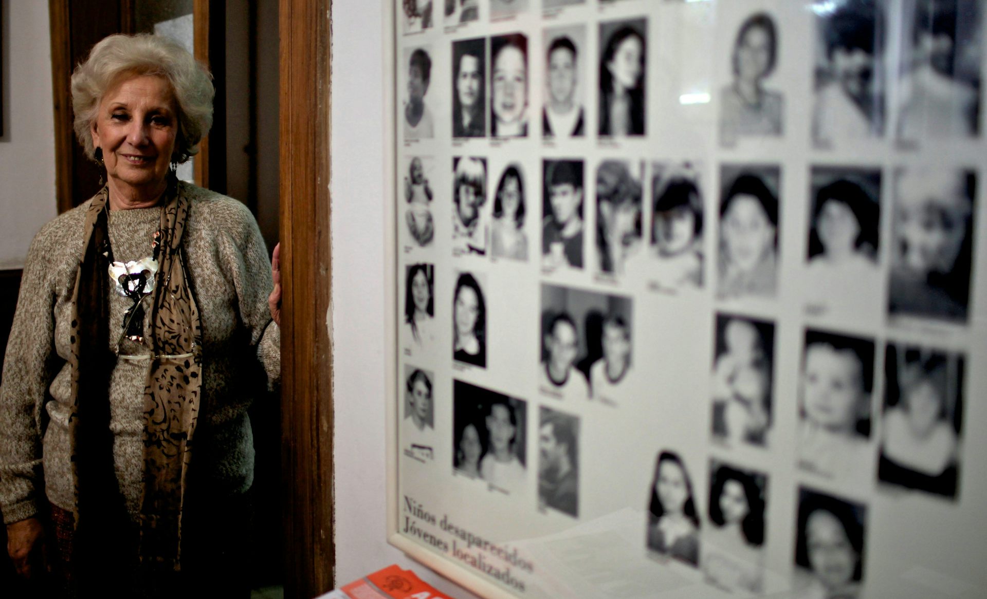 An older woman with short grey hair poses next to a poster of black and white photos
