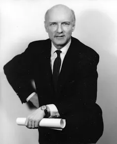 A bald man in a dark suit and tie posing with rolled up papers in his hand.