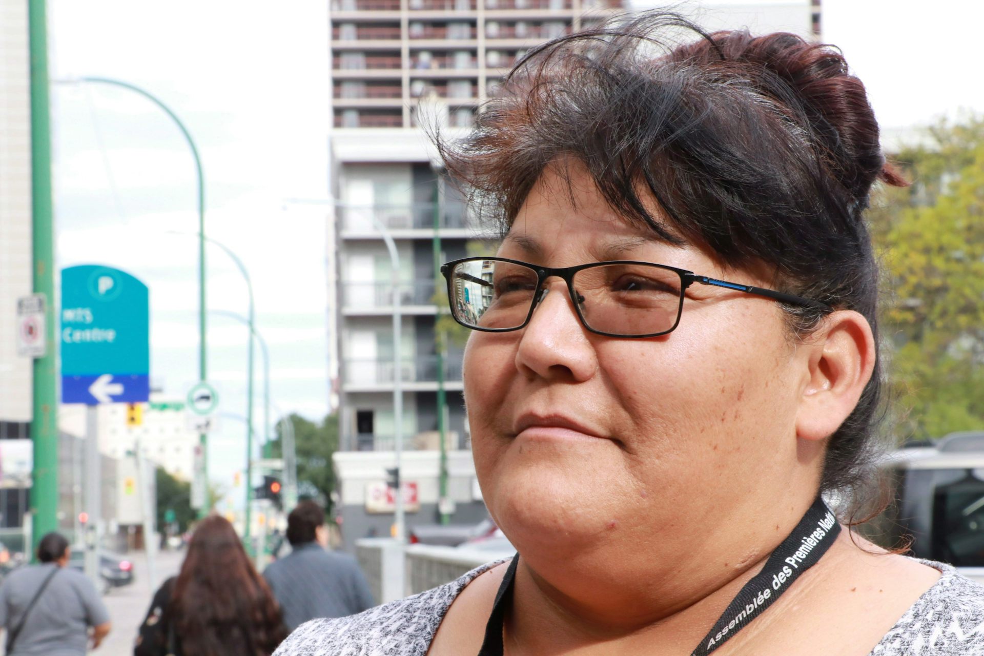 A close-up of a woman with glasses and hair up in a bun who is standing on a sidewalk.