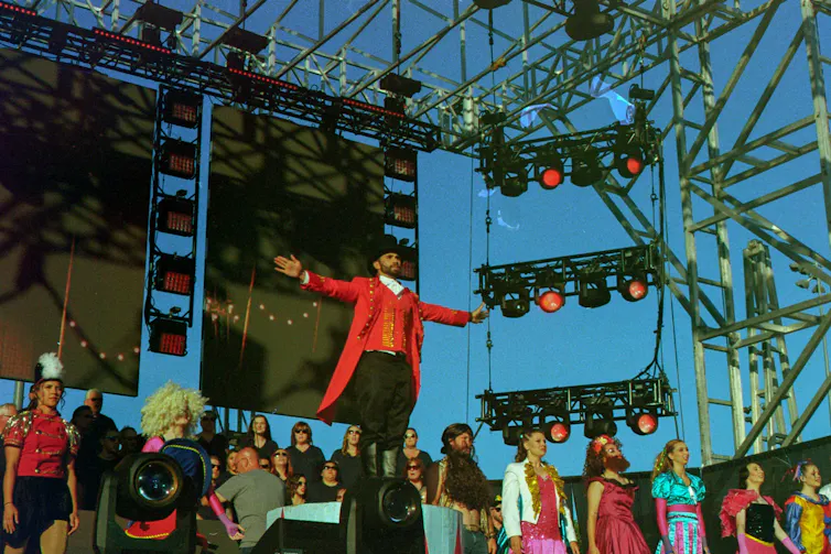 A man in a long red coat, red waistcoat, black pants and a black top hat stands with arms raised on a stage with stage lighting beside him.
