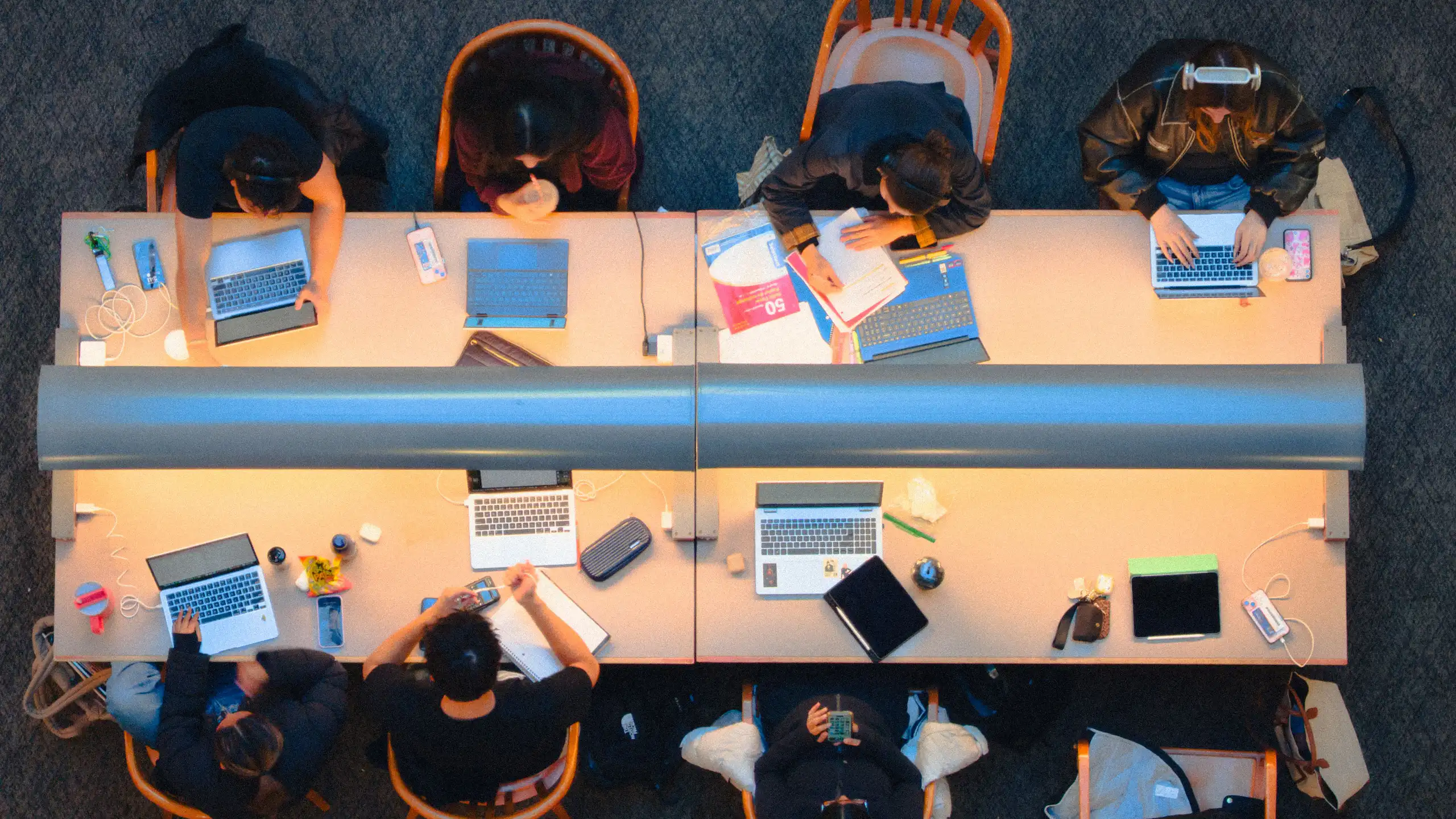 An aerial view of eight people working at a long table with computers and notebooks in a library.