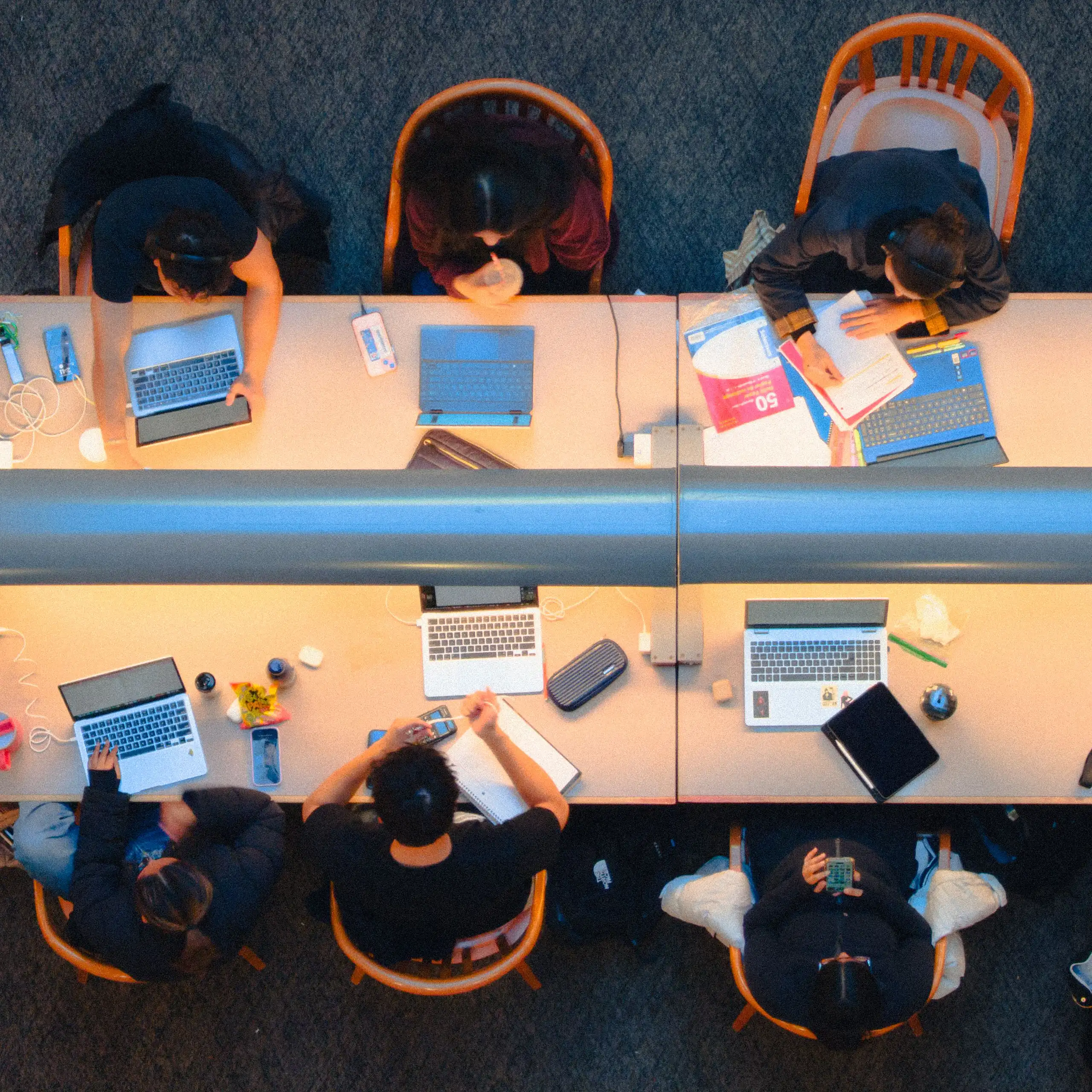 An aerial view of eight people working at a long table with computers and notebooks in a library.