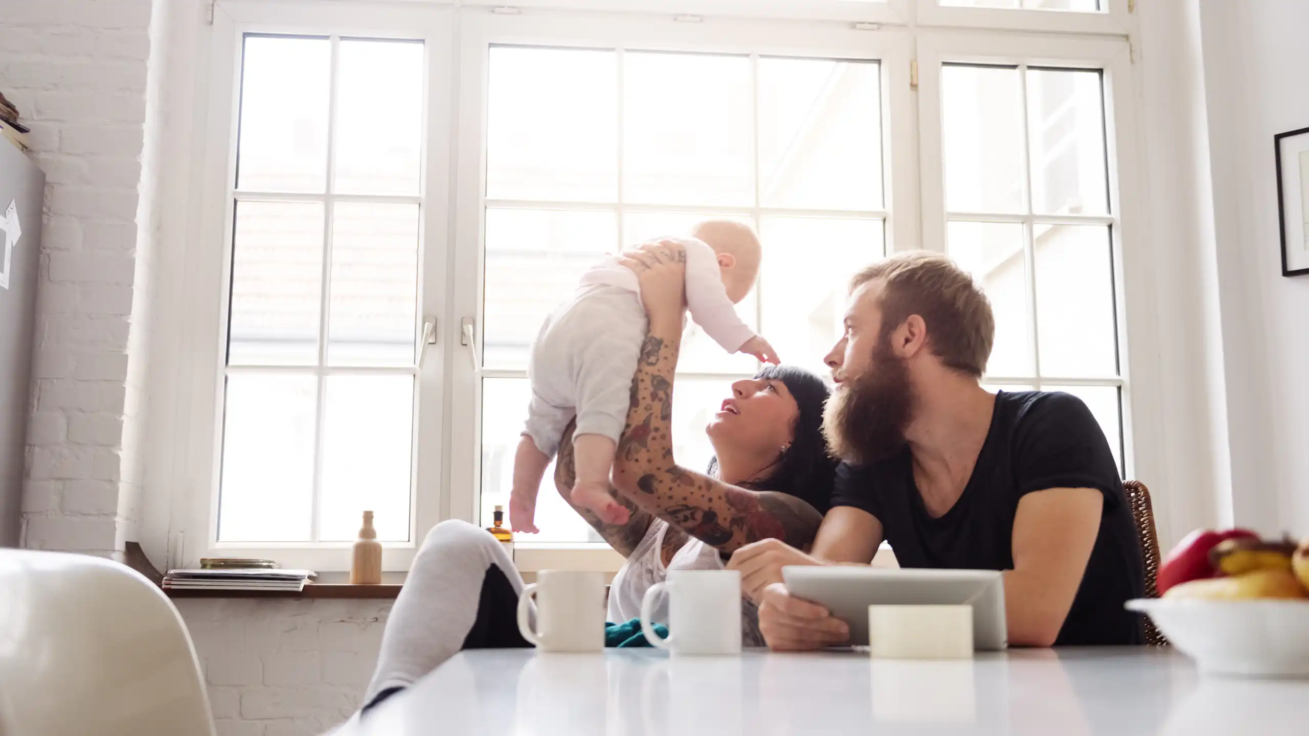 A young couple sit in front of a window holding a newborn baby